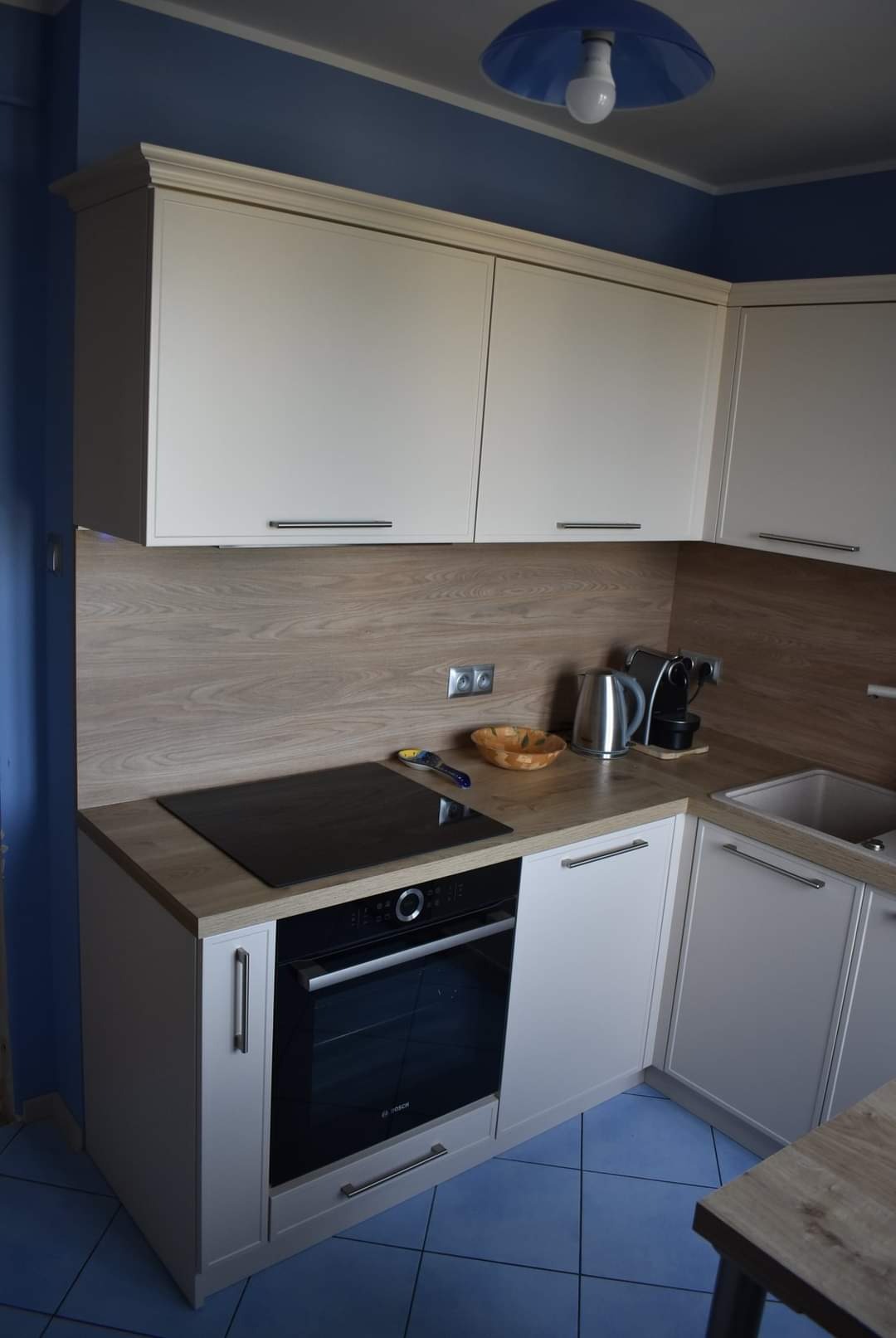 Jasne, kremowe szafki kuchenne z drewnianym blatem i niebieskimi ścianami oraz podłogą z płytek. Na blacie czarna płyta indukcyjna, piekarnik, czajnik elektryczny i ekspres do kawy.
