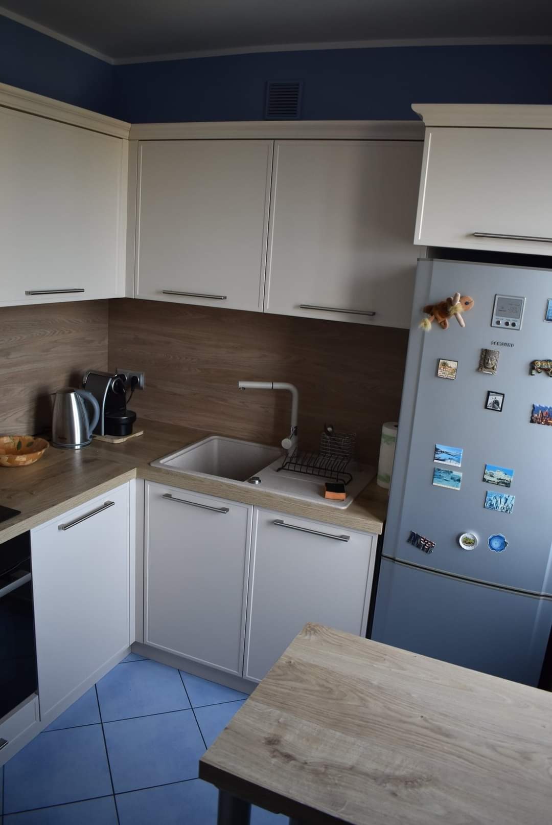 Jasna kuchnia z białymi szafkami i drewnianym blatem, niebieska podłoga i lodówka ozdobiona magnesami, widok na narożnik blatu.