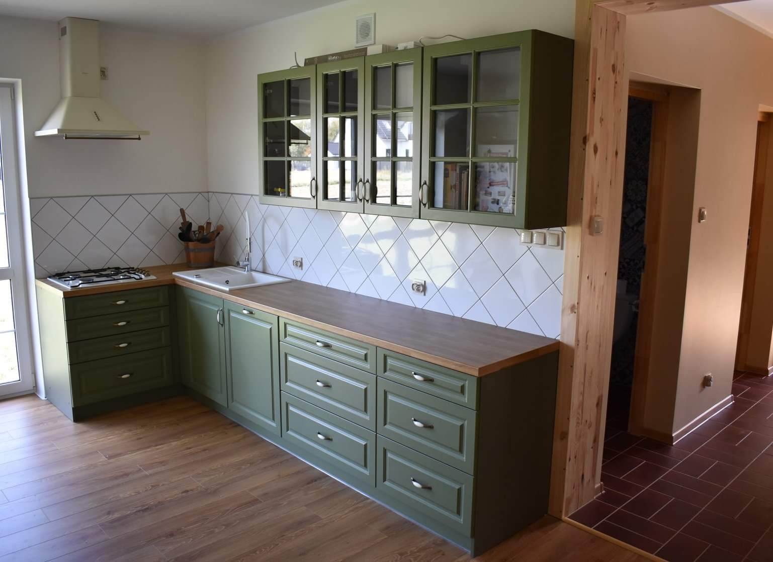 Kuchnia w odcieniach zieleni z drewnianym blatem i białymi kaflami ułożonymi w karo, widok na fragment przedpokoju z drewnianą framugą.