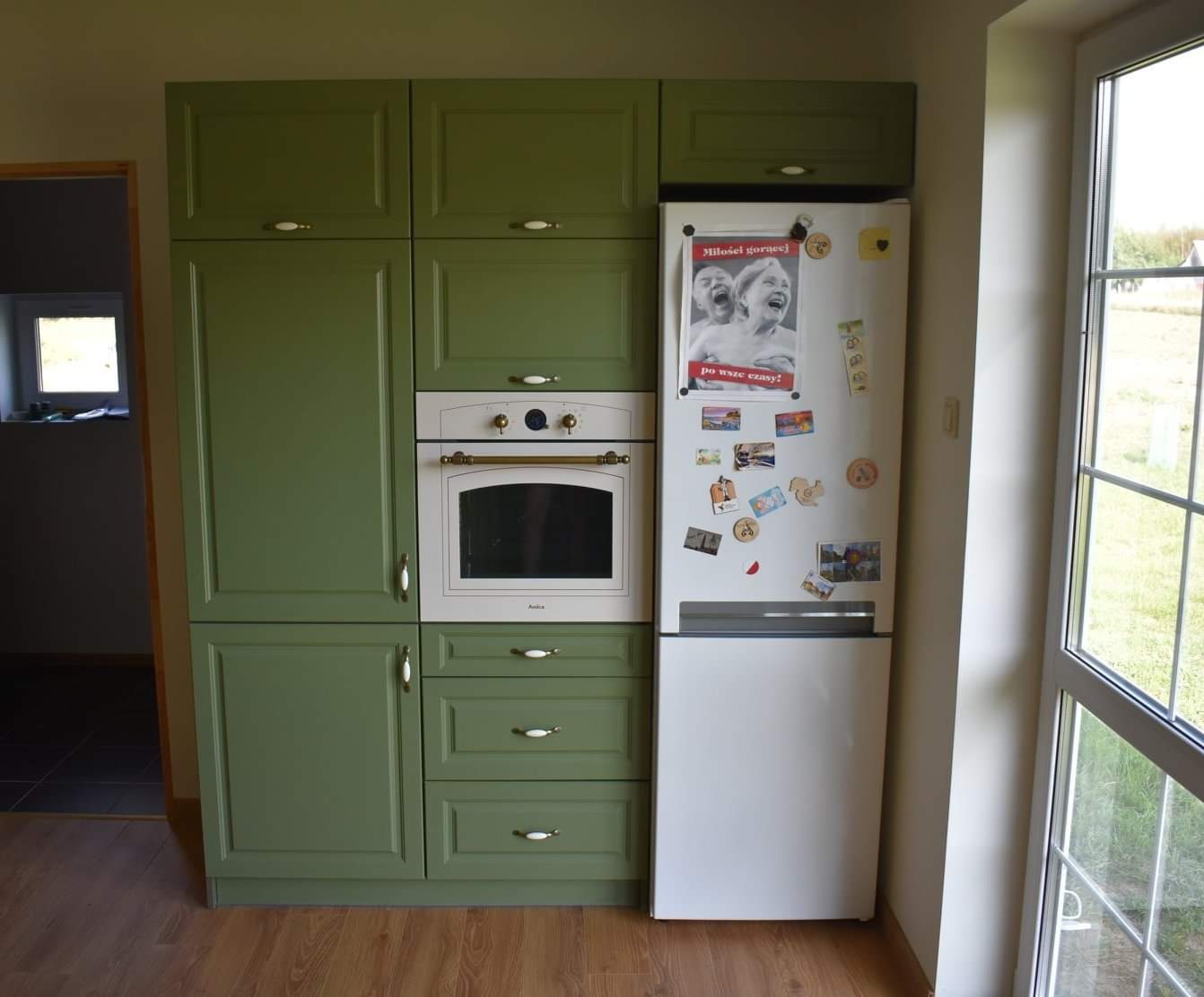 Oliwkowa zabudowa kuchenna z piekarnikiem i lodówką ozdobioną magnesami i plakatem, widok z boku na drewnianą podłogę i okno.