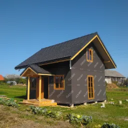 Surowy stan domu z czarną dachówką, pokrytego membraną Ruukki, z drewnianą konstrukcją ganku i okien, na tle pola z kapustą i drabiną.