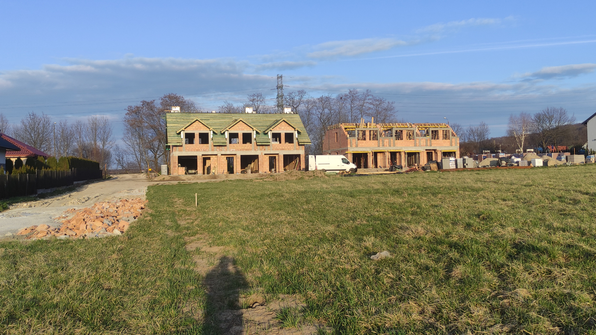 Dwa domy w stanie surowym otwartym w Łęgu Tarnowskim, jeden z zielonym dachem, drugi bez. Na pierwszym planie trawa, w tle drzewa i słupy energetyczne.