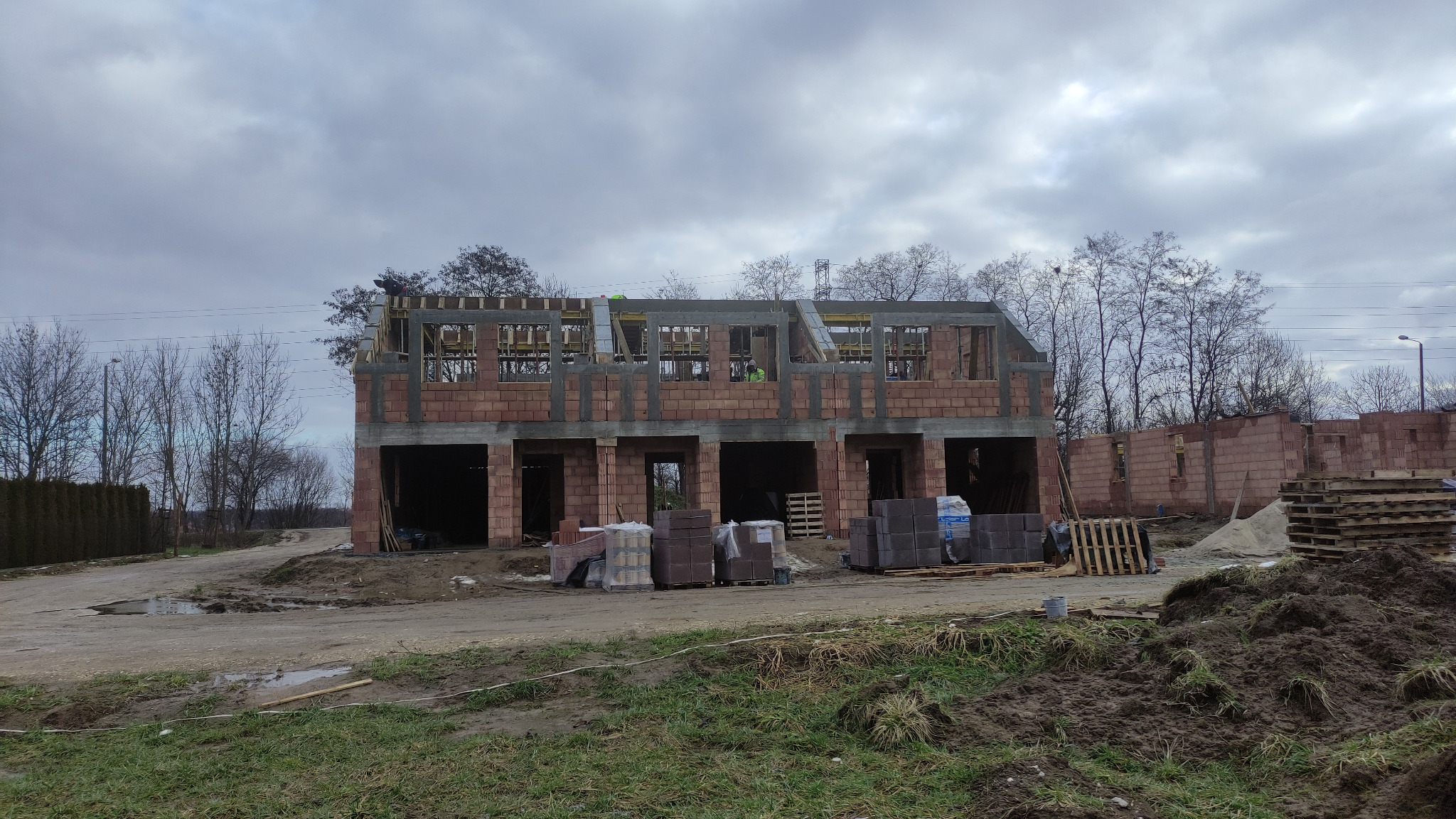 Wznoszenie surowego stanu szeregowca z czerwonej cegły. Widoczne zbrojenia i szalunki. Na pierwszym planie palety z materiałami budowlanymi i ziemia.