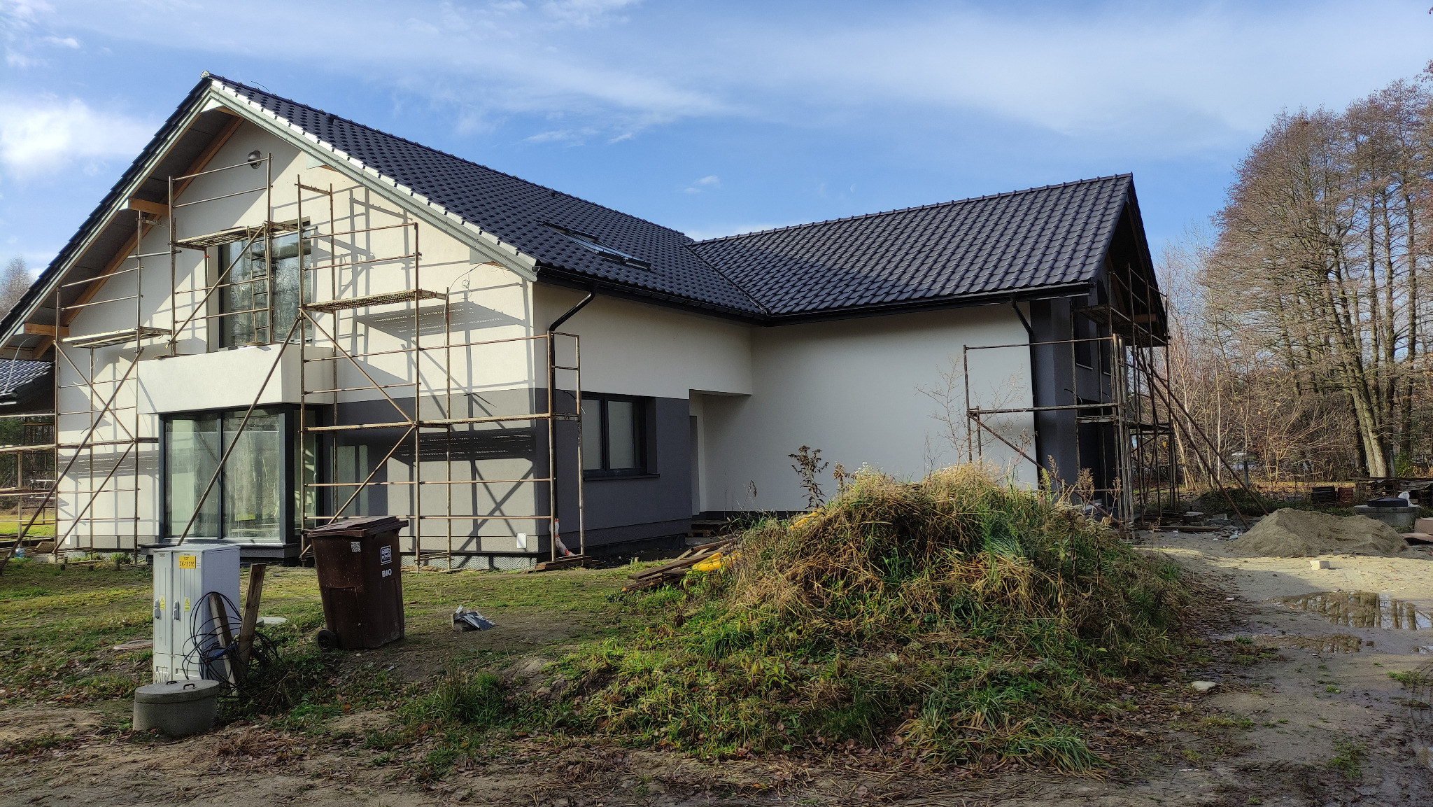 Nowoczesny dom jednorodzinny w trakcie wykończenia elewacji, z rusztowaniami i czarnym dachem, na tle błękitnego nieba i jesiennego krajobrazu.