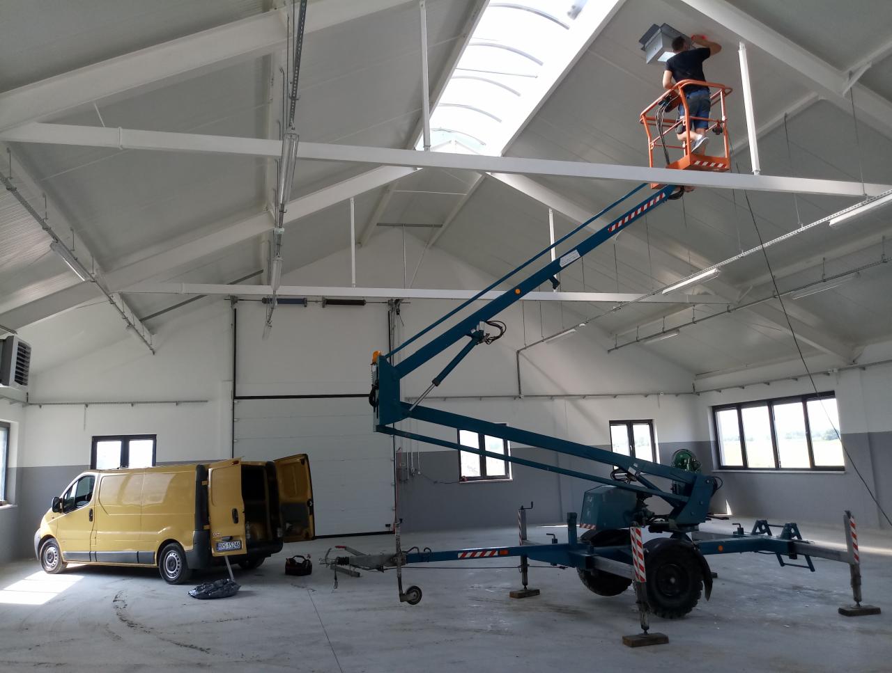Montaż systemu wentylacji w hali: pracownik na podnośniku instaluje element pod sufitem, obok żółty bus z otwartymi drzwiami i podnośnik koszowy.