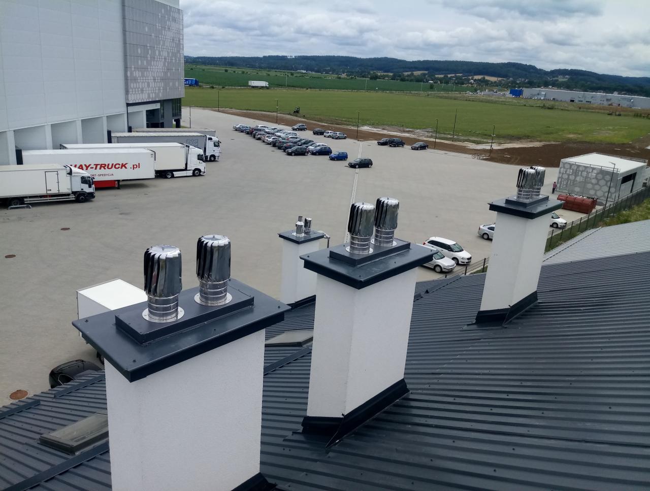 Widok z góry na cztery kominy wentylacyjne z turbinami wiatrowymi na dachu budynku przemysłowego, z parkingiem i zielonymi wzgórzami w tle.