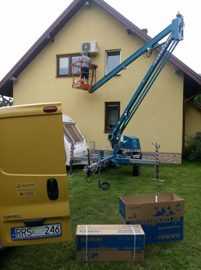 Instalacja klimatyzatora Kaisai na elewacji domu jednorodzinnego z podnośnika koszowego; w tle żółty samochód dostawczy Opel Wellmont i przyczepa kempingowa.