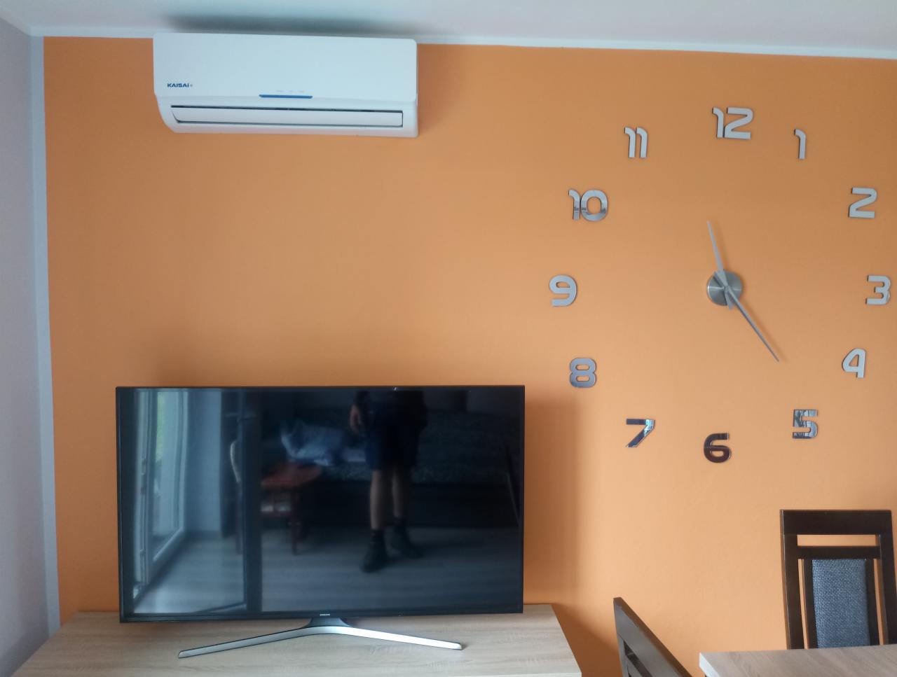 Klimatyzator marki Kaisai zamontowany w mieszkaniu w Dębicy, widoczny na pomarańczowej ścianie nad telewizorem i zegarem.