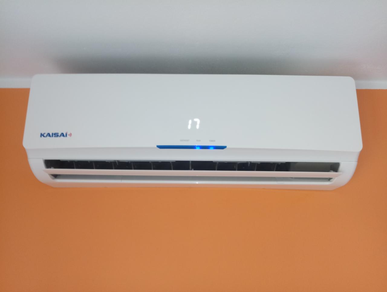 Biały klimatyzator Kaisai zamontowany na pomarańczowej ścianie, wyświetlający temperaturę 17 stopni.