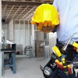 Remontowany dom w trakcie budowy z widocznym pracownikiem budowlanym trzymającym kask i młotek, z pasem narzędziowym, na tle surowych ścian i drewnianej konstrukcji stropu.