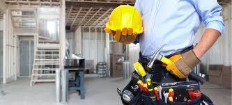 Remontowany dom w trakcie budowy z widocznym pracownikiem budowlanym trzymającym kask i młotek, z pasem narzędziowym, na tle surowych ścian i drewnianej konstrukcji stropu.