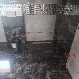 Usługi Remontowo-Budowlane Jarosław Drozd - Łazienka w trakcie układania płytek: szare płytki imitujące marmur na podłodze i ścianach, poziomowane pomarańczowymi klipsami, widoczny fragment okna dachowego i pojemnik na klej.