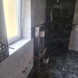 Usługi Remontowo-Budowlane Jarosław Drozd - Układanie płytek w łazience: szare i czarne marmurowe płytki na ścianach i podłodze, widoczne krzyżyki dystansowe i pojemnik z narzędziami.