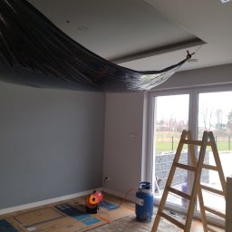 Usługi Remontowo-Budowlane Jarosław Drozd - Przygotowanie do prac malarskich: pomieszczenie zabezpieczone folią od góry, kartonem na podłodze, nagrzewnica i butla gazowa w rogu, drabina oparta o okno.