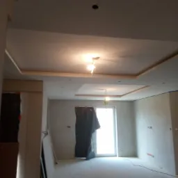 Wnętrze w trakcie remontu: sufit z podwieszaną konstrukcją z płyt kartonowo-gipsowych, ściany przygotowane pod malowanie, okno zasłonięte folią ochronną, widoczne przewody elektryczne.