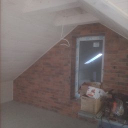Usługi Remontowo-Budowlane Jarosław Drozd - Poddasze w trakcie wykańczania: surowa cegła, okno PCV, odeskowana skośna ściana i fragment narzędzi na stole warsztatowym.