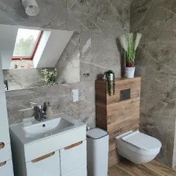 Nowoczesna łazienka z marmurowymi płytkami, wiszącą toaletą z drewnianą obudową, białą szafką pod umywalką i drewnianą podłogą. W lustrze odbija się okno dachowe. Dekoracyjne rośliny dodają wnętrzu...