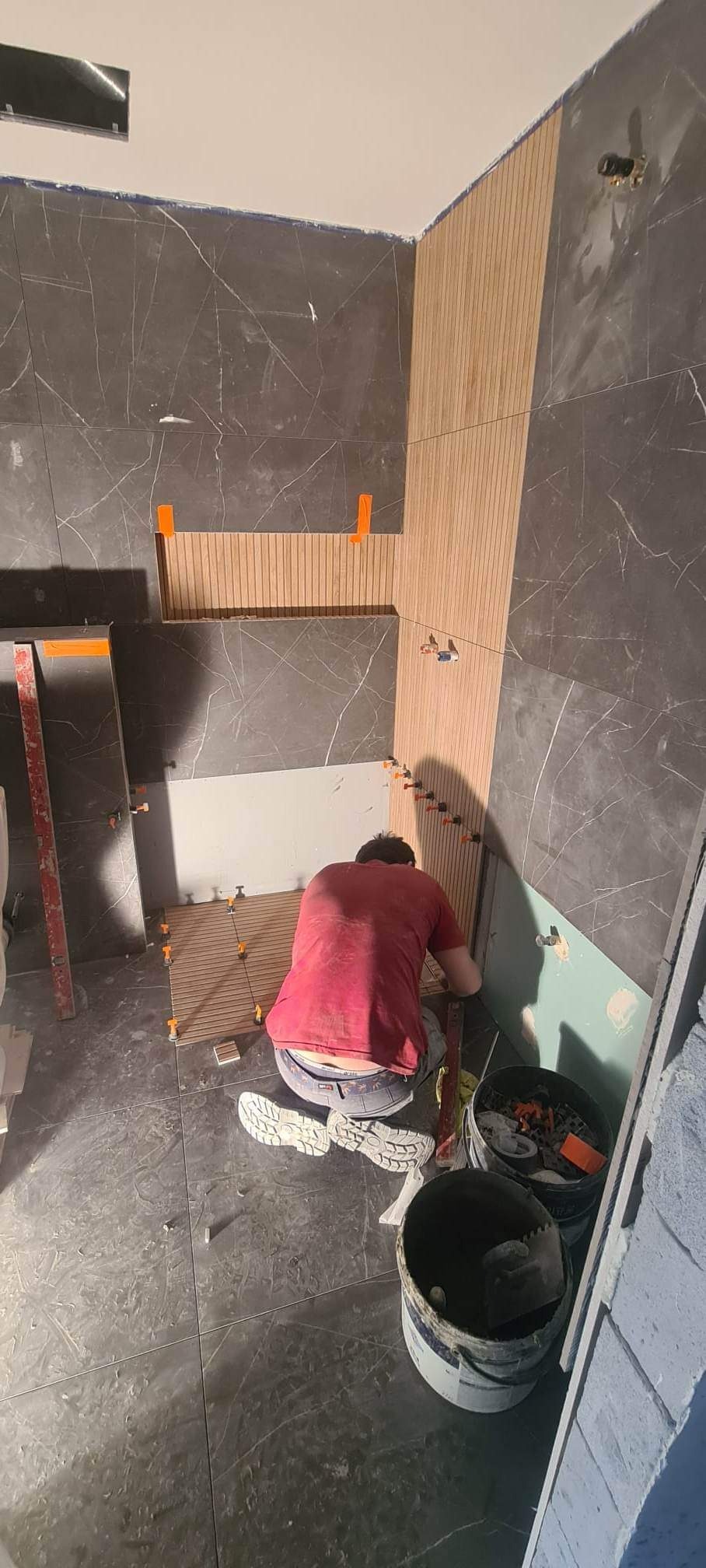 Glazurnik w trakcie układania drewnopodobnych płytek na podłodze i ścianie w łazience z elementami szarego marmuru, widoczne narzędzia i poziomica.