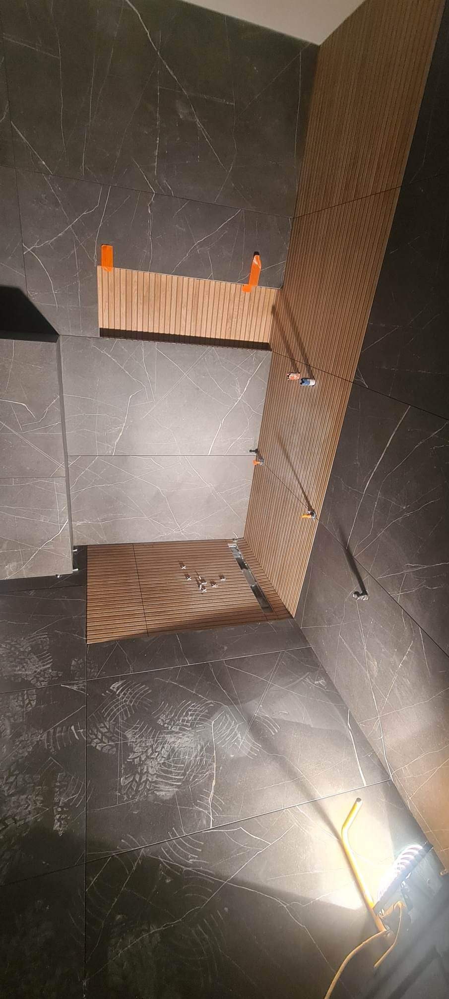 Montaż drewnopodobnych paneli ściennych i podłogowych w łazience, widoczne klipsy poziomujące, ciemnoszara płytka imitująca marmur na ścianach, ślady butów na podłodze i lampa robocza.