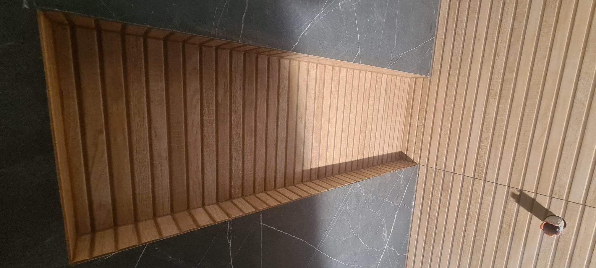 Wnęka w ścianie wyłożona drewnianymi lamelami, przylegająca do ściany z płytek imitujących ciemny marmur.