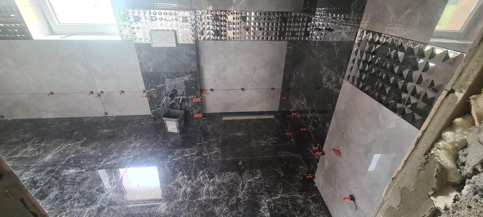 Łazienka w trakcie układania płytek: szare płytki imitujące marmur na podłodze i ścianach, poziomowane pomarańczowymi klipsami, widoczny fragment okna dachowego i pojemnik na klej.