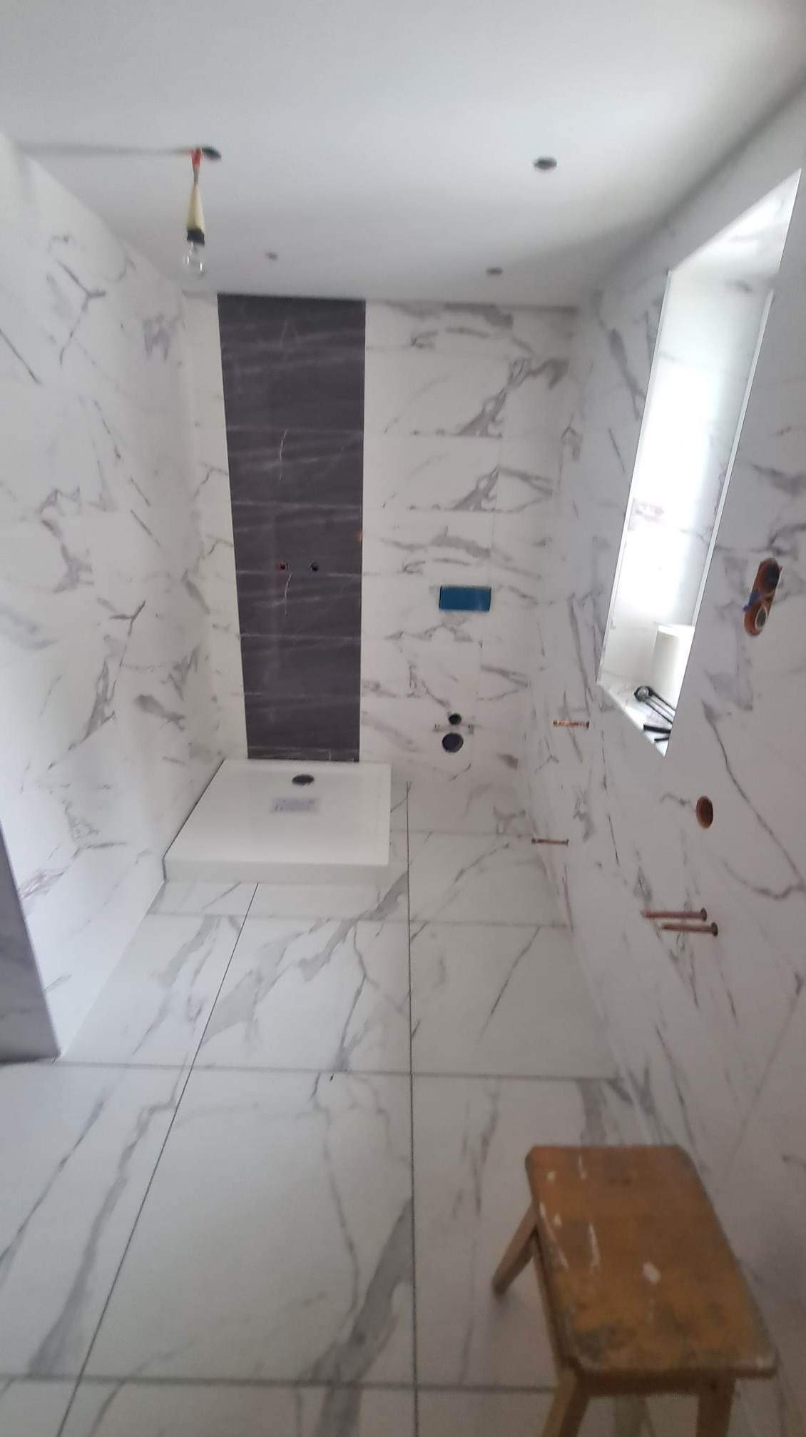 Wnętrze łazienki w trakcie wykańczania z białymi marmurkowymi płytkami, ciemnoszary pas płytek na ścianie, biały brodzik i drewniany stołek w rogu.