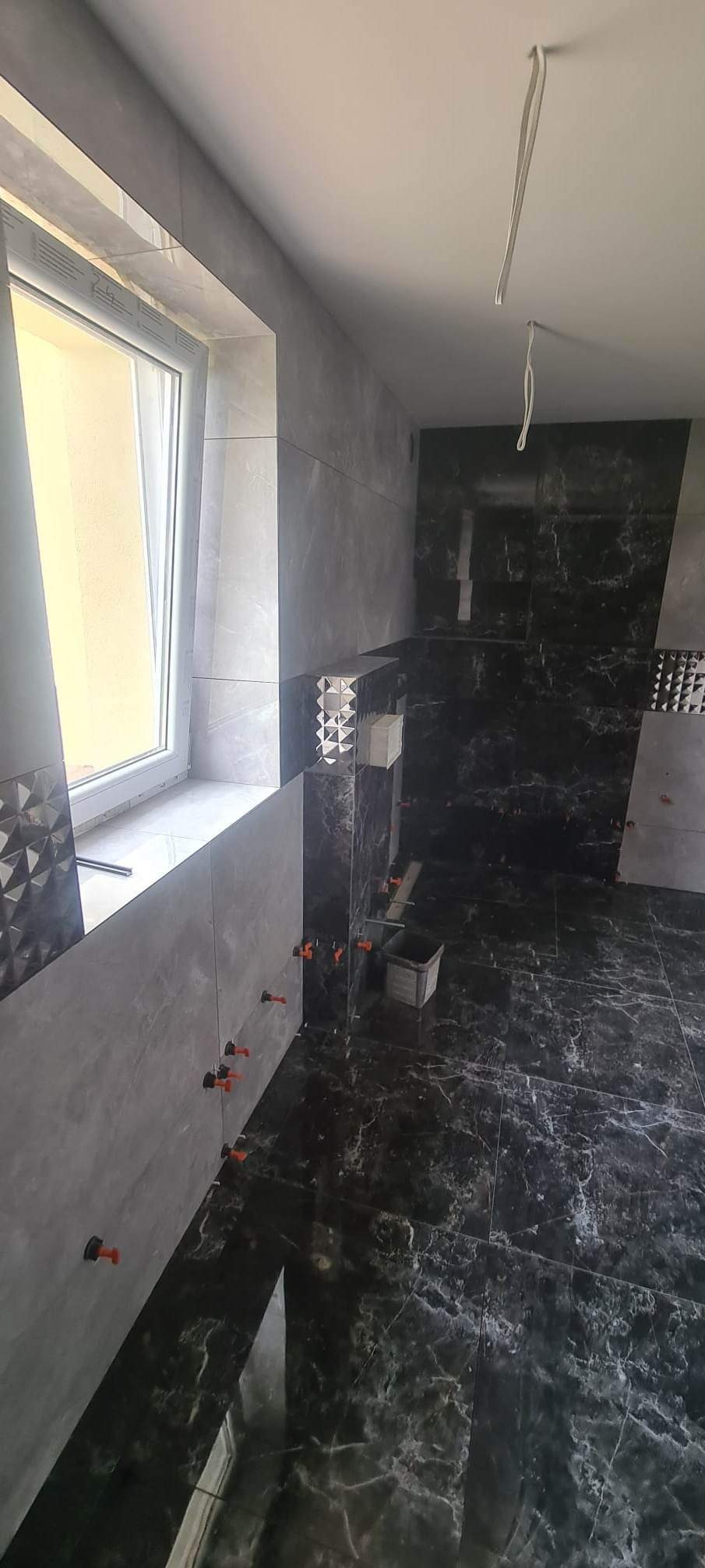 Układanie płytek w łazience: szare i czarne marmurowe płytki na ścianach i podłodze, widoczne krzyżyki dystansowe i pojemnik z narzędziami.