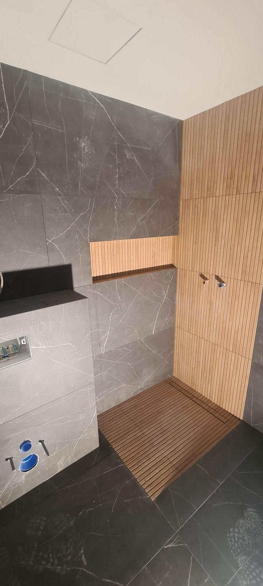 Nowoczesna kabina prysznicowa z szarymi płytkami imitującymi marmur, drewnianymi lamelami na ścianie i podłogą z drewnianych listew; widoczne elementy instalacji wodnej.