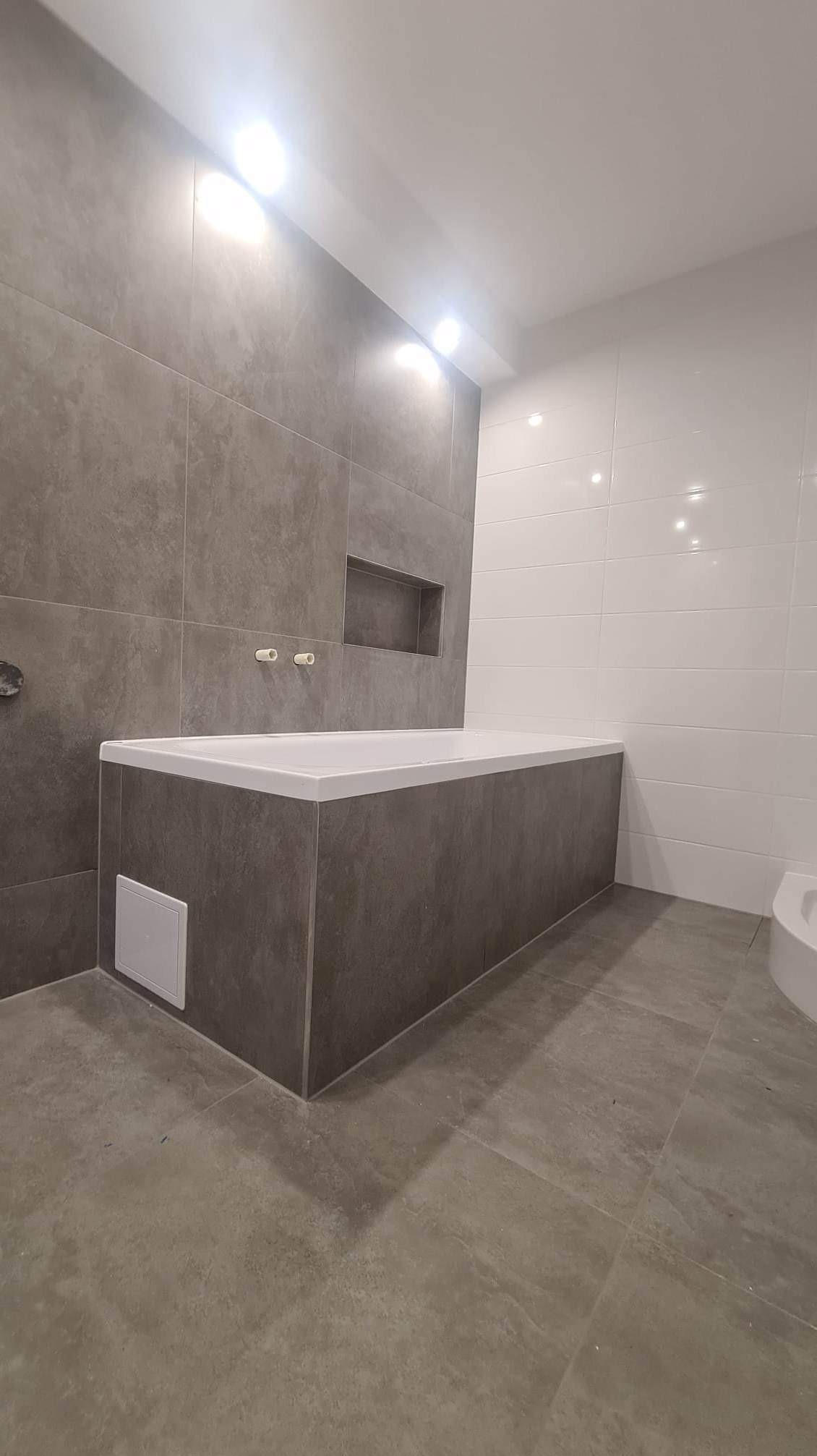 Wykończona łazienka z zabudowaną wanną w szarych płytkach, wnęka w ścianie nad wanną, białe płytki na części ściany, szara podłoga z płytek.