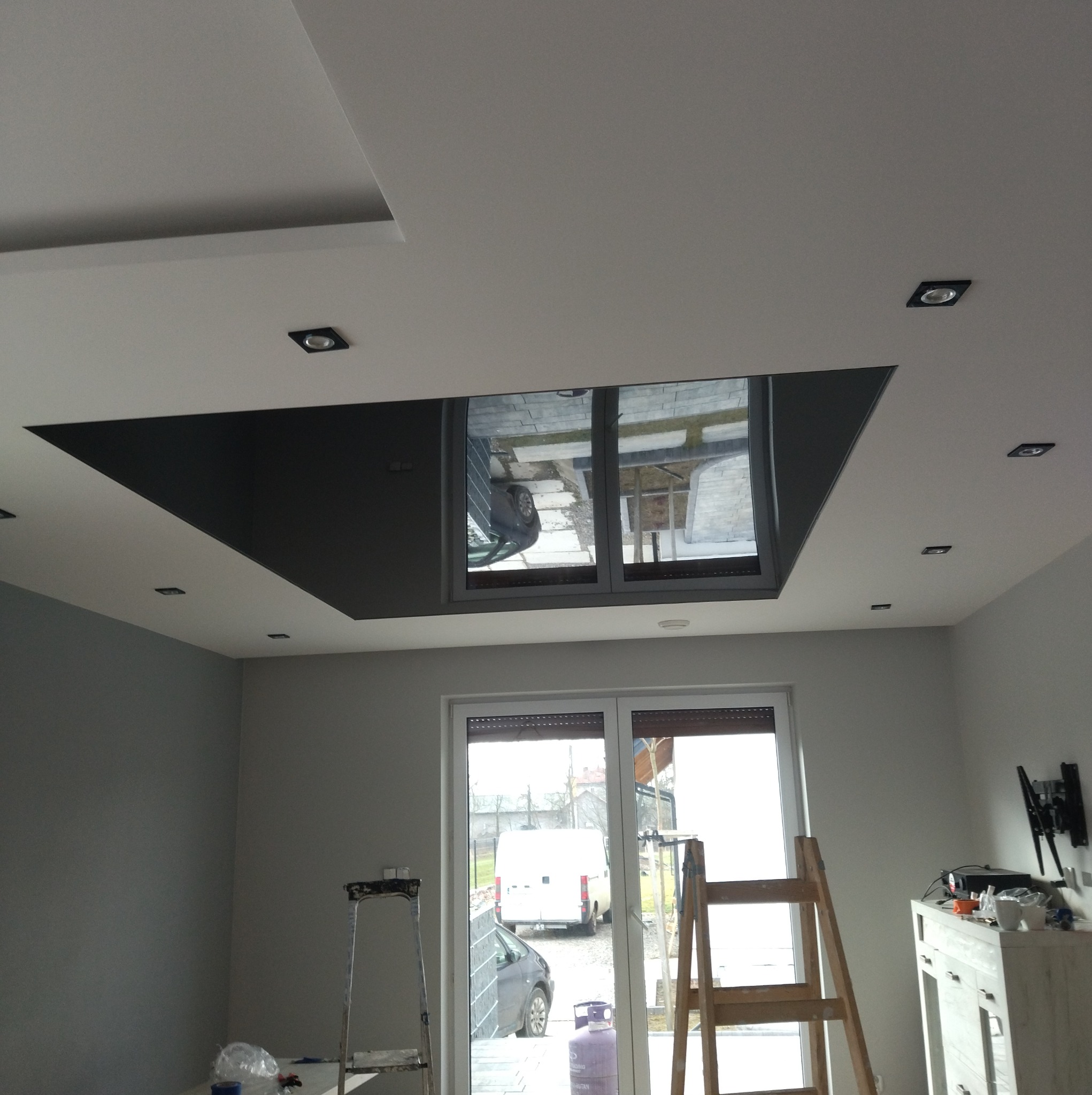 Pokój po remoncie z pomalowanymi ścianami na szaro, z oknem balkonowym, schodami malarskimi, sufitem podwieszanym z czarnym wstawkami i oświetleniem punktowym.