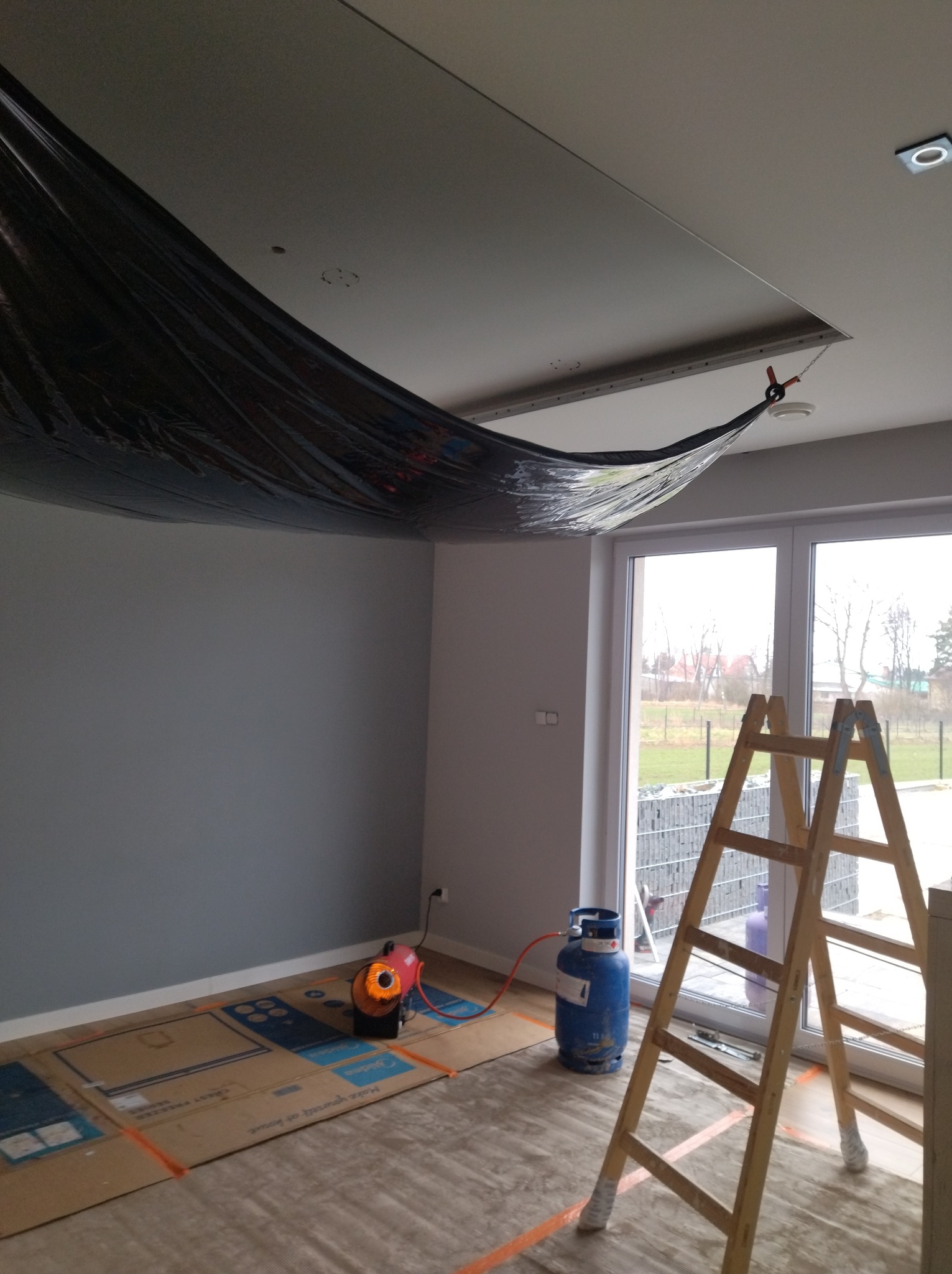 Przygotowanie do prac malarskich: pomieszczenie zabezpieczone folią od góry, kartonem na podłodze, nagrzewnica i butla gazowa w rogu, drabina oparta o okno.