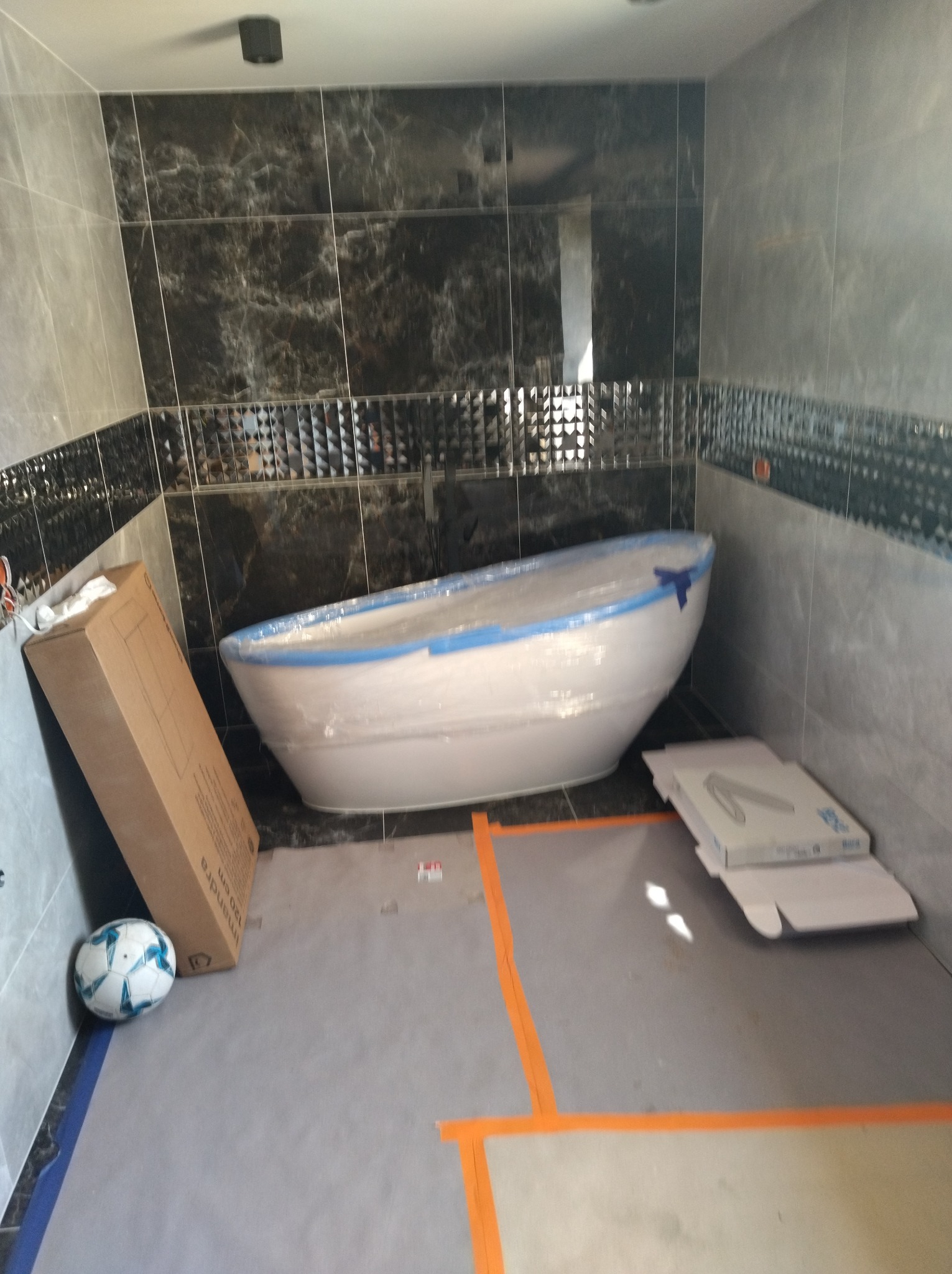 Nowa wanna wolnostojąca w folii ochronnej, ustawiona w łazience z szarymi i czarnymi płytkami, widoczny karton po wannie i piłka do nogi.