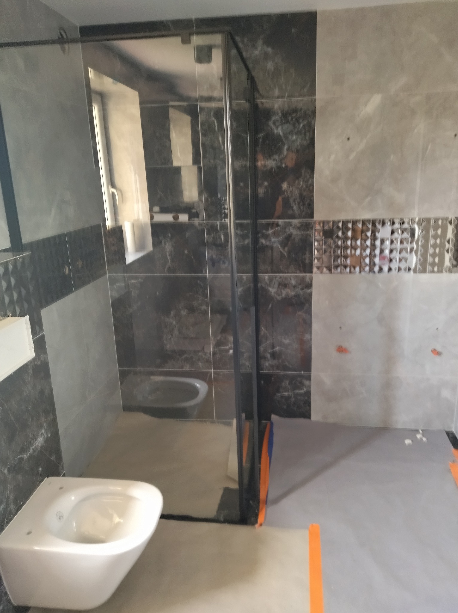 Montaż podwieszanej miski WC w łazience z szarymi płytkami imitującymi marmur, widoczna kabina prysznicowa z czarnymi profilami i zabezpieczona podłoga.