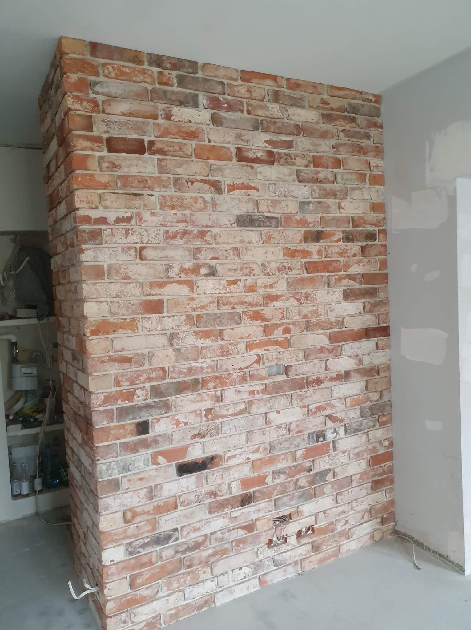 Ściana wyłożona dekoracyjną cegłą w odcieniach czerwieni i bieli, widoczne niedokończone prace wykończeniowe przy podłodze i ścianie obok.