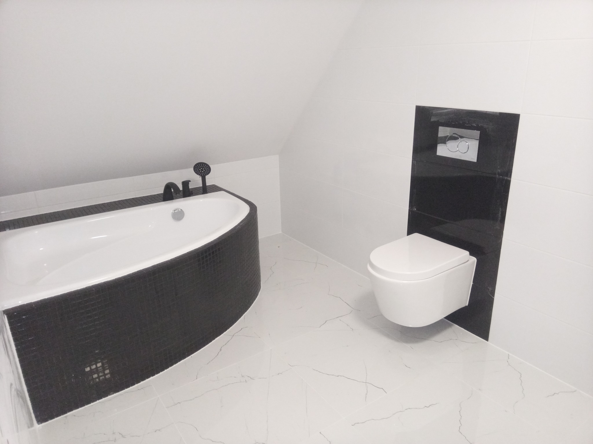 Nowoczesna łazienka na poddaszu z białą wanną obudowaną czarną mozaiką, wiszącą toaletą i marmurową podłogą.