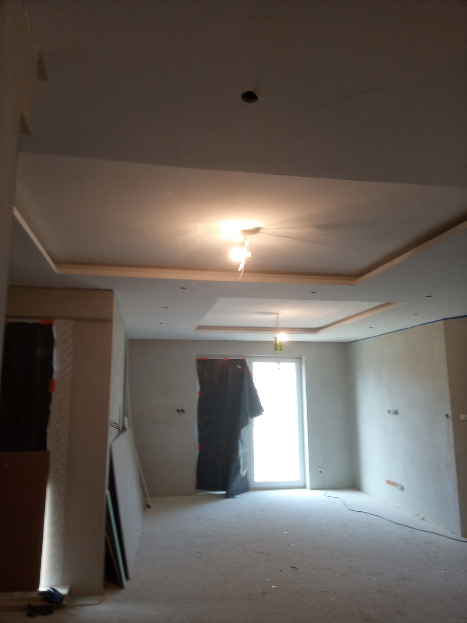 Wnętrze w trakcie remontu: sufit z podwieszaną konstrukcją z płyt kartonowo-gipsowych, ściany przygotowane pod malowanie, okno zasłonięte folią ochronną, widoczne przewody elektryczne.