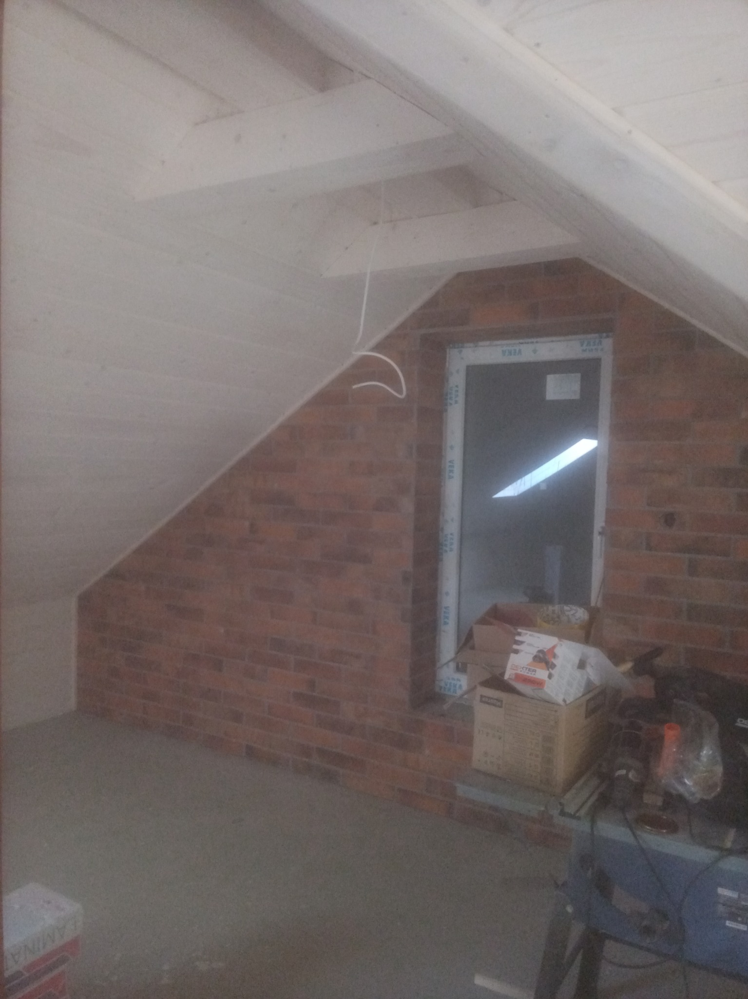 Poddasze w trakcie wykańczania: surowa cegła, okno PCV, odeskowana skośna ściana i fragment narzędzi na stole warsztatowym.