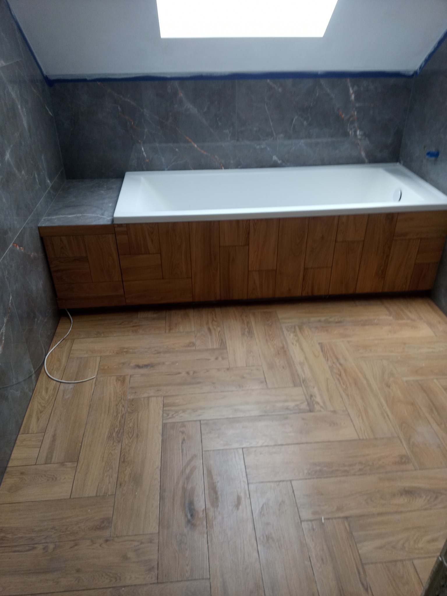 Nowoczesna łazienka z wanną obudowaną drewnopodobną płytą, szare marmurowe płytki na ścianach i podłoga ułożona w jodełkę.