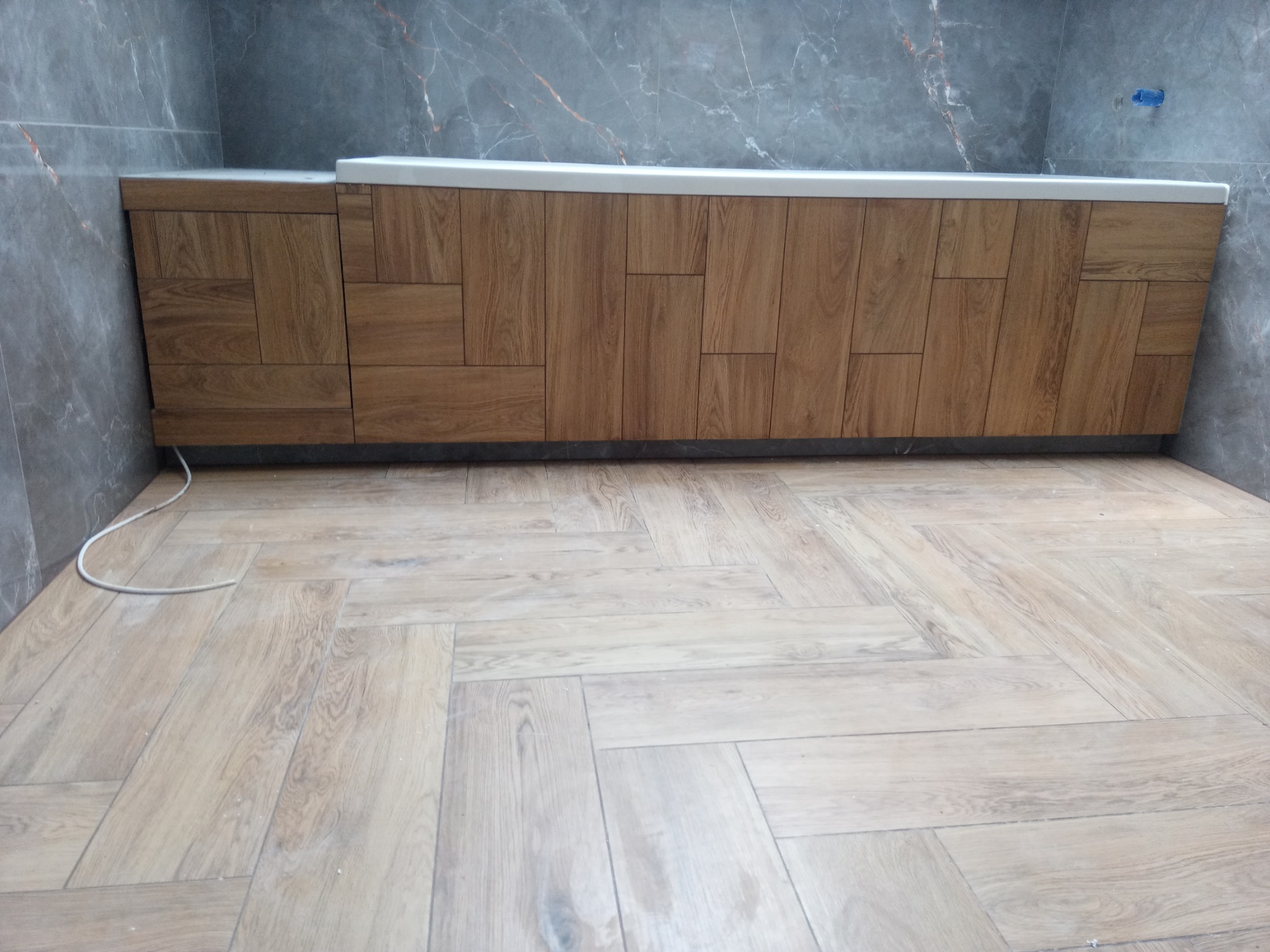 Wykończona łazienka z drewnopodobną zabudową wanny i podłogą ułożoną w jodełkę, szare marmurowe płytki na ścianach.