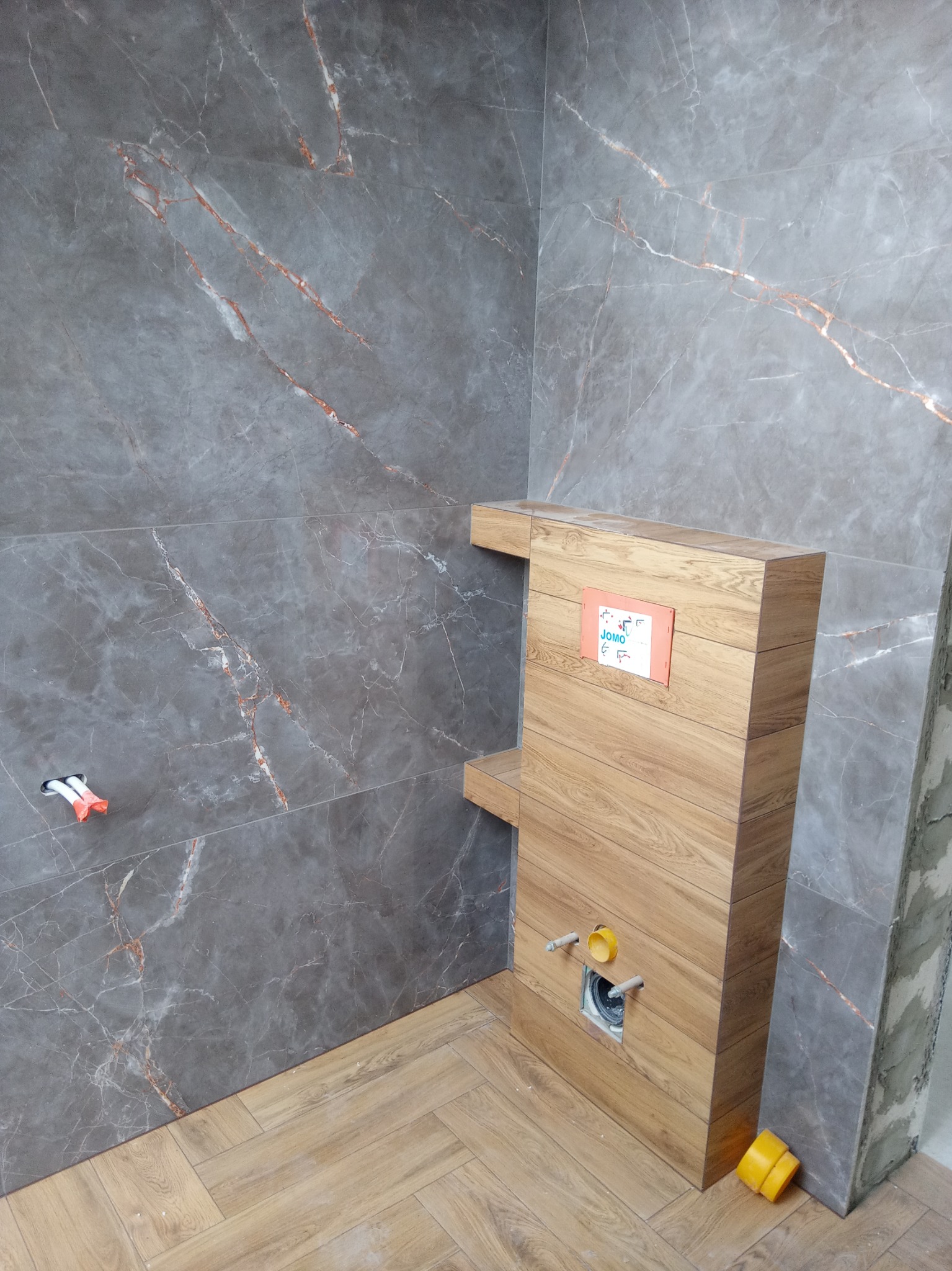 Montaż podtynkowego stelaża WC z obudową imitującą drewno na tle ściany wyłożonej szarymi płytkami imitującymi marmur, widoczne przyłącza wodne i kanalizacyjne, podłoga wyłożona parkietem w jodełkę.