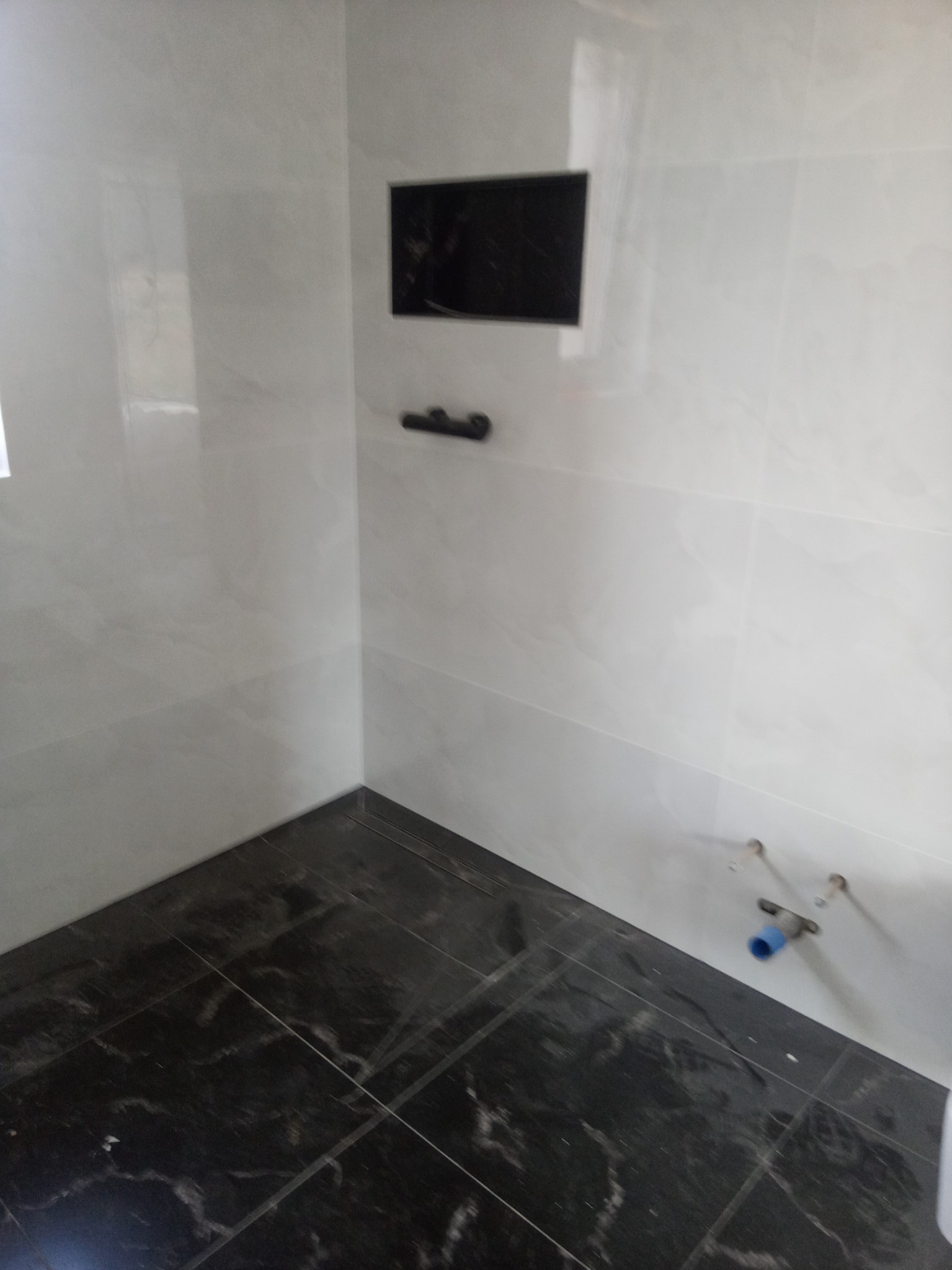 Wykończona łazienka z czarną podłogą z płytek imitujących marmur, białe ściany z wnęką i czarnym uchwytem na ręcznik, widoczne przyłącza hydrauliczne.