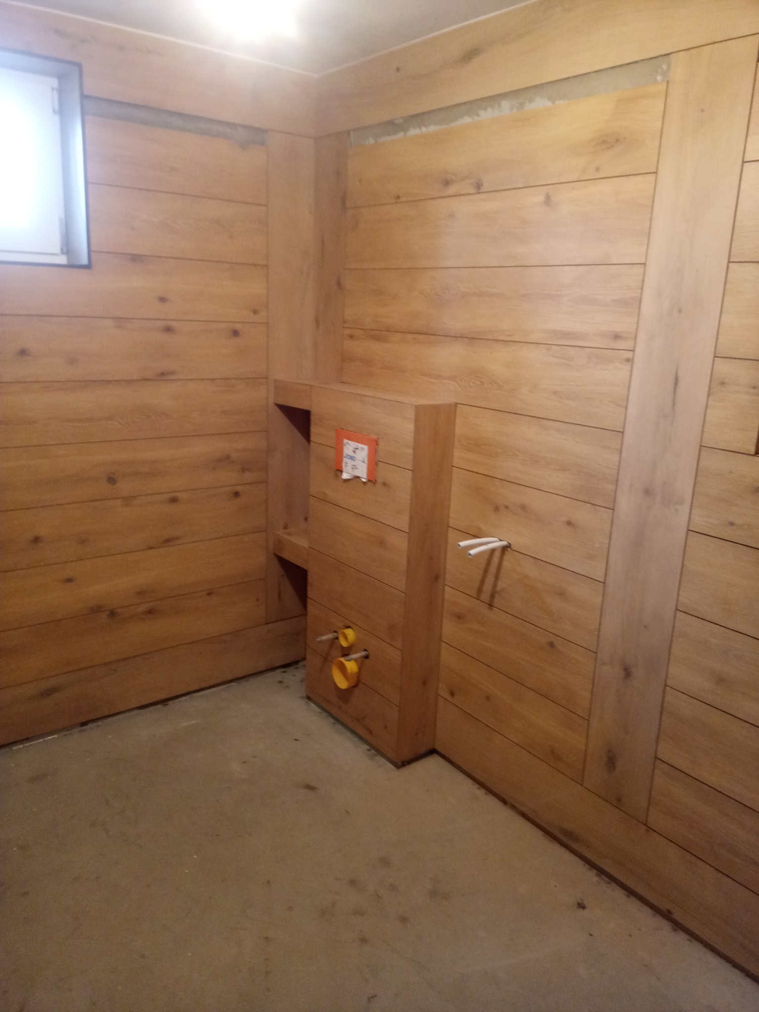 Łazienka w trakcie wykańczania, ściany wyłożone panelami drewnopodobnymi, widoczne przyłącza hydrauliczne do toalety podtynkowej, surowa podłoga.