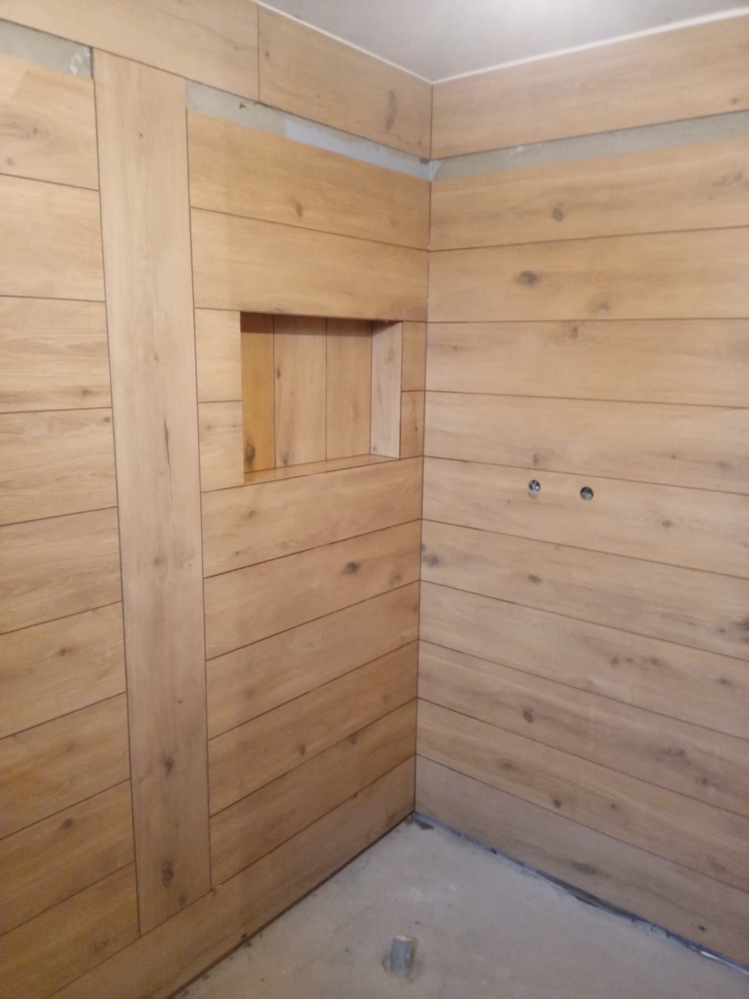 Wykończona ściana łazienki z płytek imitujących drewno, z wnęką na kosmetyki i widocznymi otworami na instalację elektryczną.