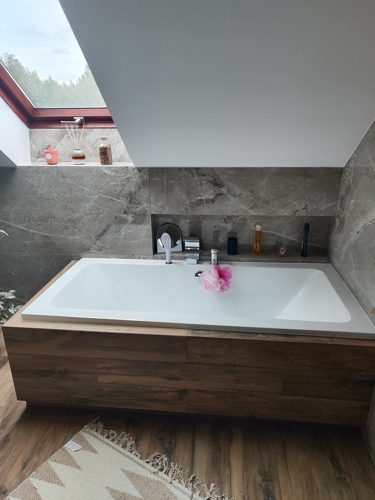 Nowoczesna łazienka na poddaszu z wanną obudowaną drewnem, marmurowe płytki ścienne i okno dachowe wpuszczające światło dzienne.