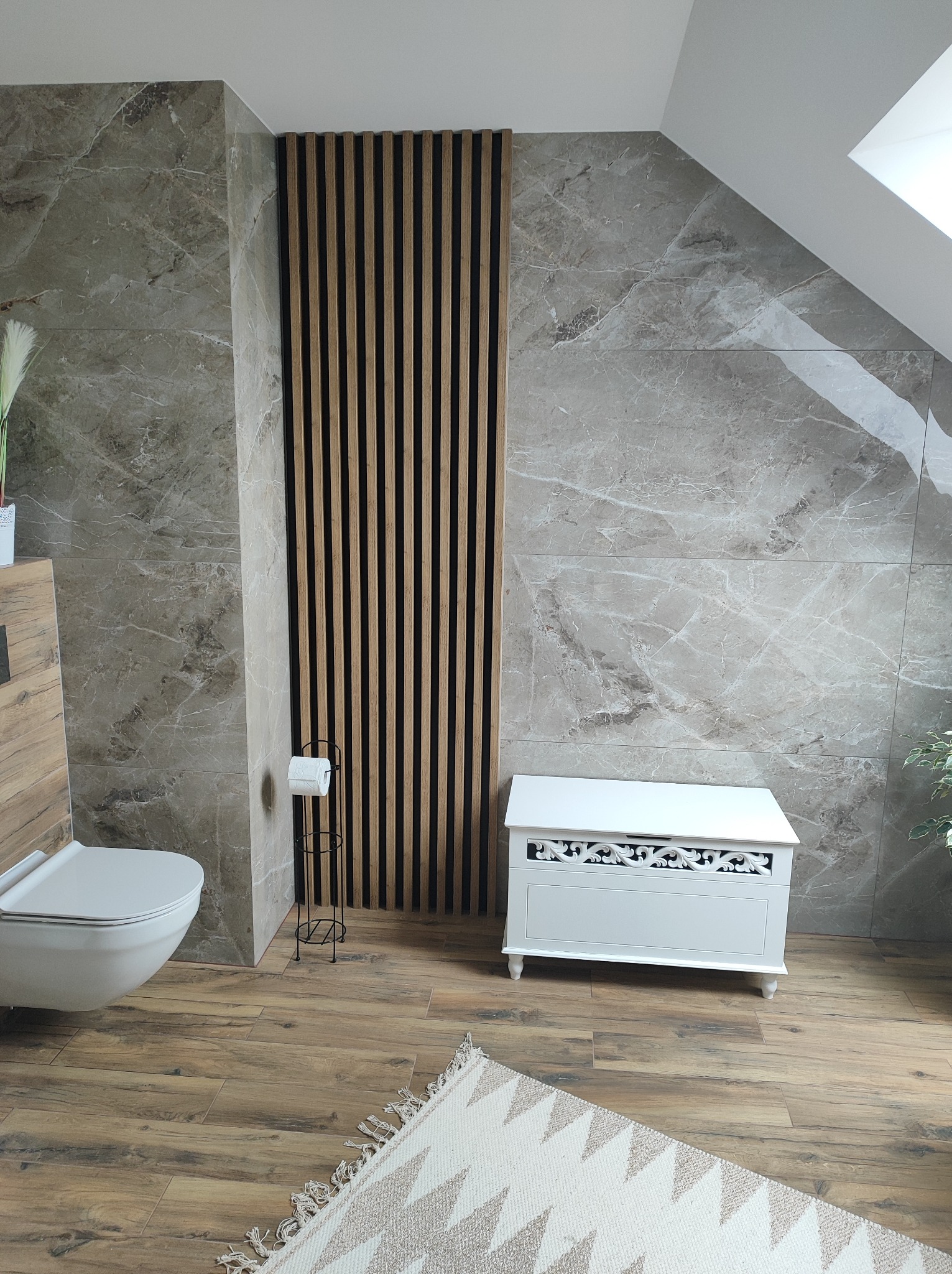 Wykończona łazienka w odcieniach szarości i brązu z drewnianymi lamelami dekoracyjnymi, toaleta podwieszana, biała komoda z ornamentami oraz dywanik z frędzlami.