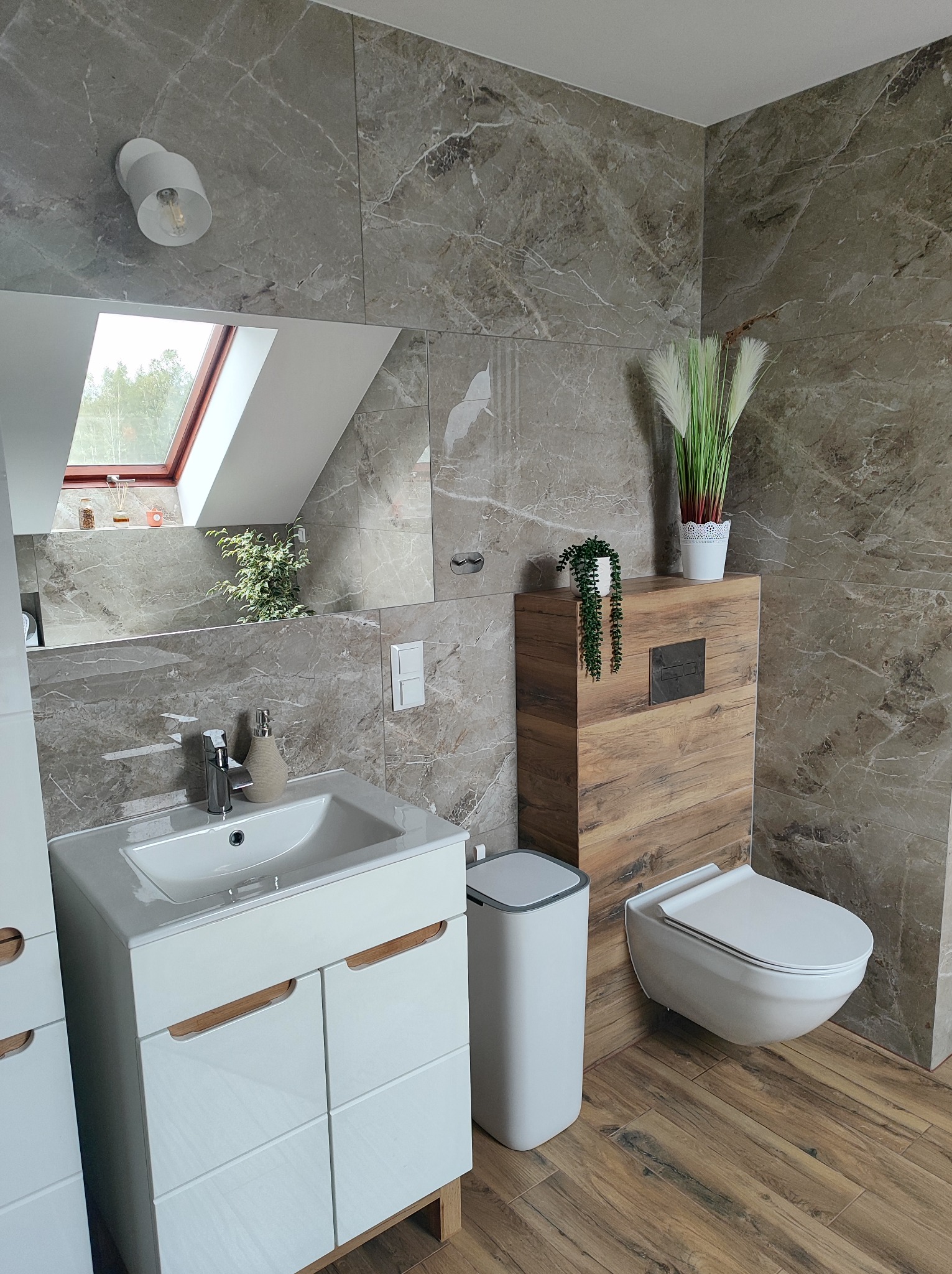 Nowoczesna łazienka z marmurowymi płytkami, wiszącą toaletą z drewnianą obudową, białą szafką pod umywalką i drewnianą podłogą. W lustrze odbija się okno dachowe. Dekoracyjne rośliny dodają wnętrzu...