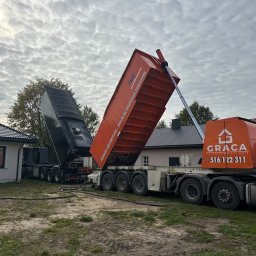 GRACA Kamil Graczyński - Dwa samochody ciężarowe z otwartymi naczepami, z których wylewana jest wylewka anhydrytowa. Widoczne węże i budynki w tle. Jeden z samochodów oznaczony logiem firmy GRACA.