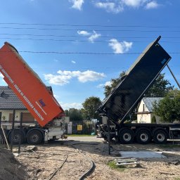 GRACA Kamil Graczyński - Dwa samochody ciężarowe z otwartymi skrzyniami, przygotowane do wylewania posadzki na budowie. Widoczne węże i teren przygotowany pod wylewkę. Błękitne niebo w tle.