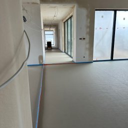 GRACA Kamil Graczyński - Gładka, szara posadzka w długim korytarzu z dużymi oknami, zabezpieczona niebieską taśmą. Widoczne kable i surowe ściany w trakcie wykończenia.
