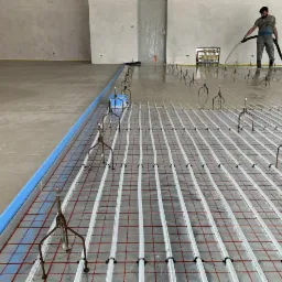 Instalacja ogrzewania podłogowego zalewana płynną wylewką, widoczne rury grzewcze, metalowe uchwyty i pracownik rozprowadzający wylewkę z węża.